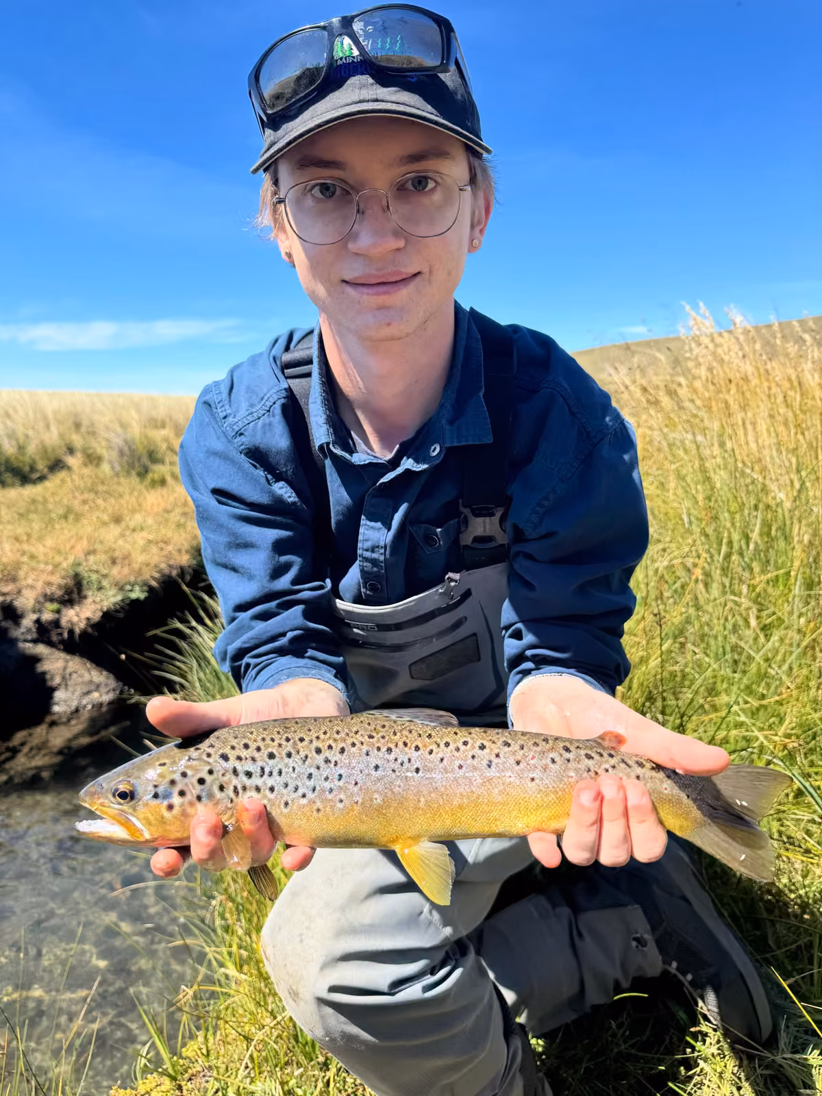 A smiling angler in a dark cap crouches on a grassy bank beside the river holding a brown trout with black spots and a yellow belly, blue sky and dry hills behind, Ñirehuao River, Coyhaique, Chilean Patagonia