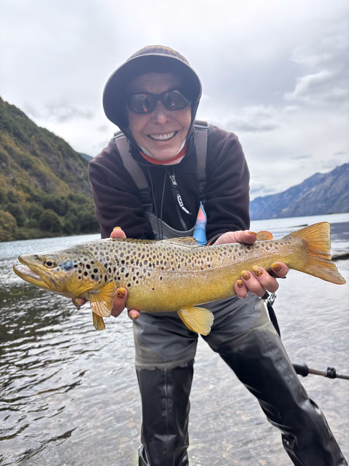 An angler in a dark hat and sunglasses stands in shallow water holding a golden brown trout with dark spots, a forested mountainside behind, Coyhaique, Chilean Patagonia