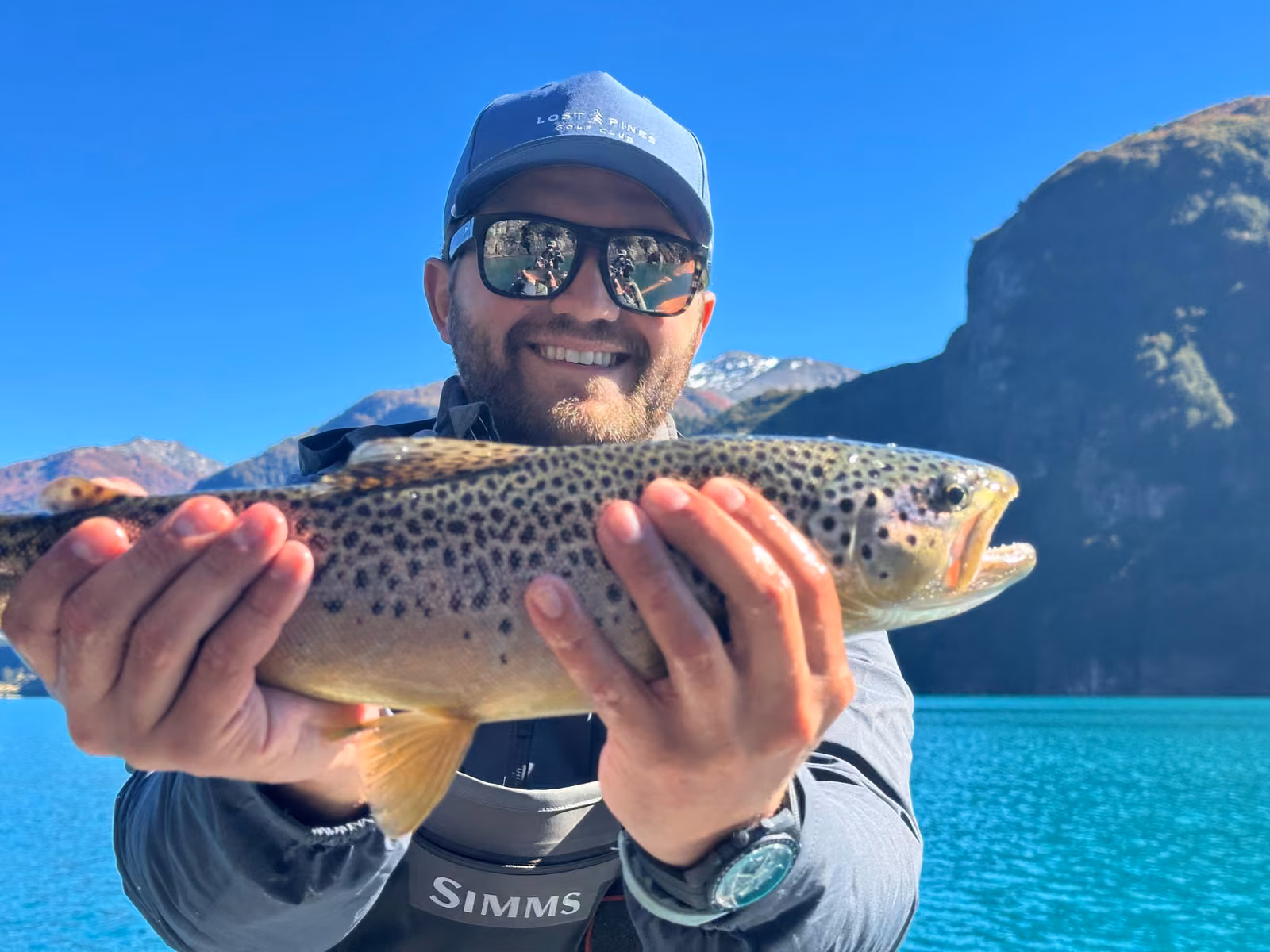 A smiling bearded angler in a blue cap and mirrored sunglasses holds a spotted brown trout in both hands, a turquoise lake and a rock wall behind, Coyhaique, Chilean Patagonia