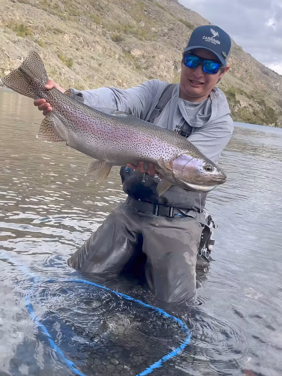 An angler in a cap stands in lake water up to the thighs and lifts a second large rainbow trout, with a dry hillside behind, Chilean Patagonia