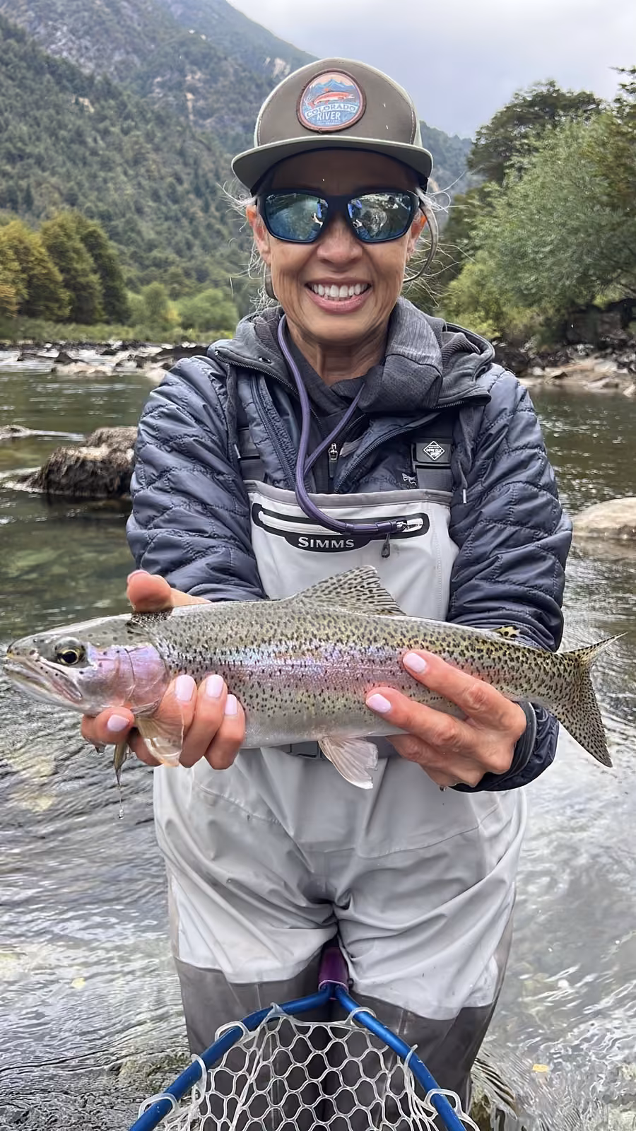A smiling angler in a cap and mirrored sunglasses holds a rainbow trout with a pink band along its flank, a forested river bank behind, Coyhaique, Chilean Patagonia
