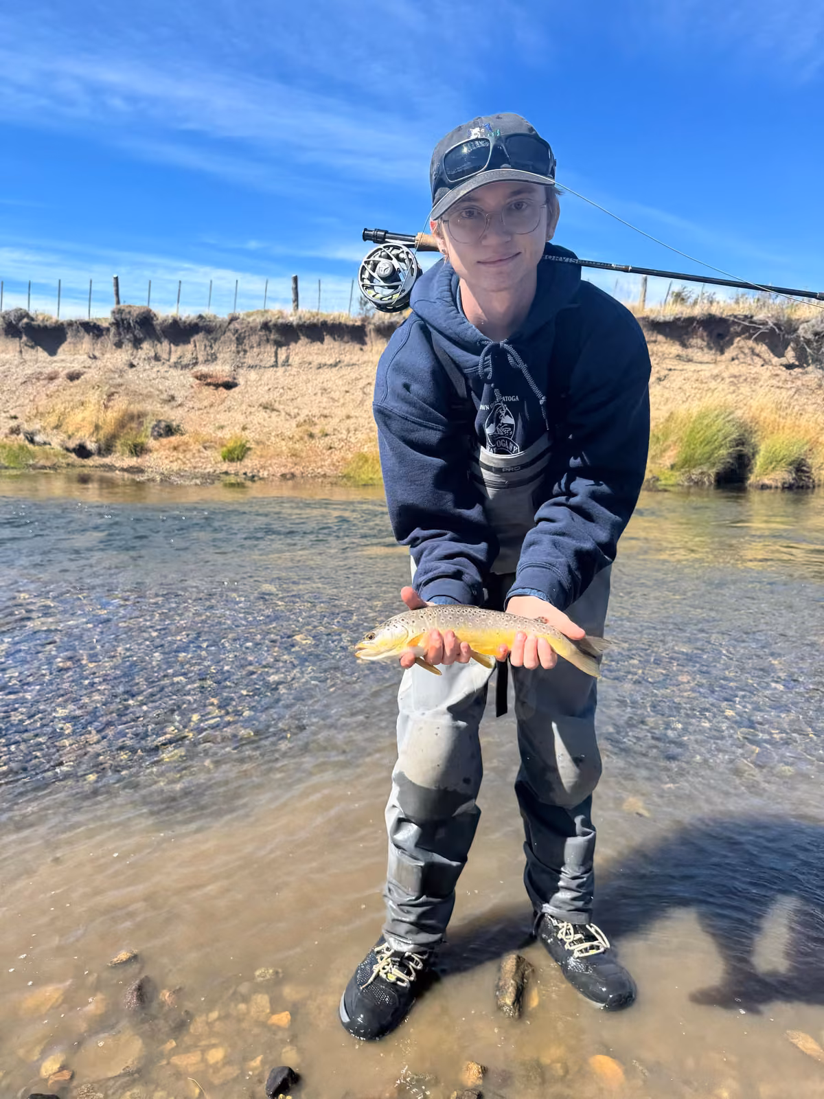 An angler in waders and a navy hoodie stands in a shallow, stony river run holding a brown trout, a fly rod over one shoulder and a dry, eroded bank behind, Ñirehuao River, Coyhaique, Chilean Patagonia