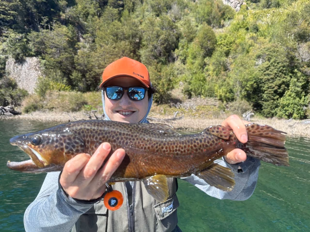 A smiling angler holds a heavily spotted wild brown trout over clear green lake water near Coyhaique, Chilean Patagonia