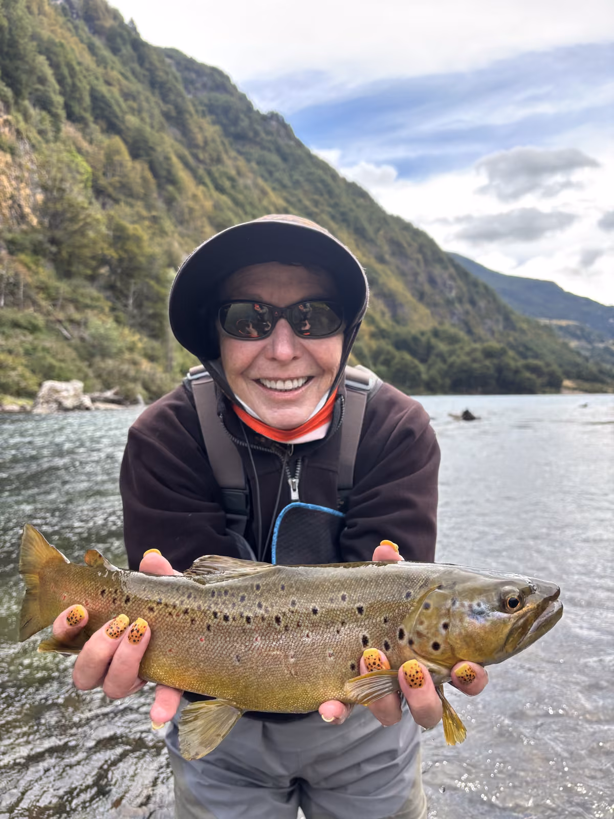 An angler in a dark hat and sunglasses stands in clear water holding a spotted brown trout, forested slopes behind, Coyhaique, Chilean Patagonia