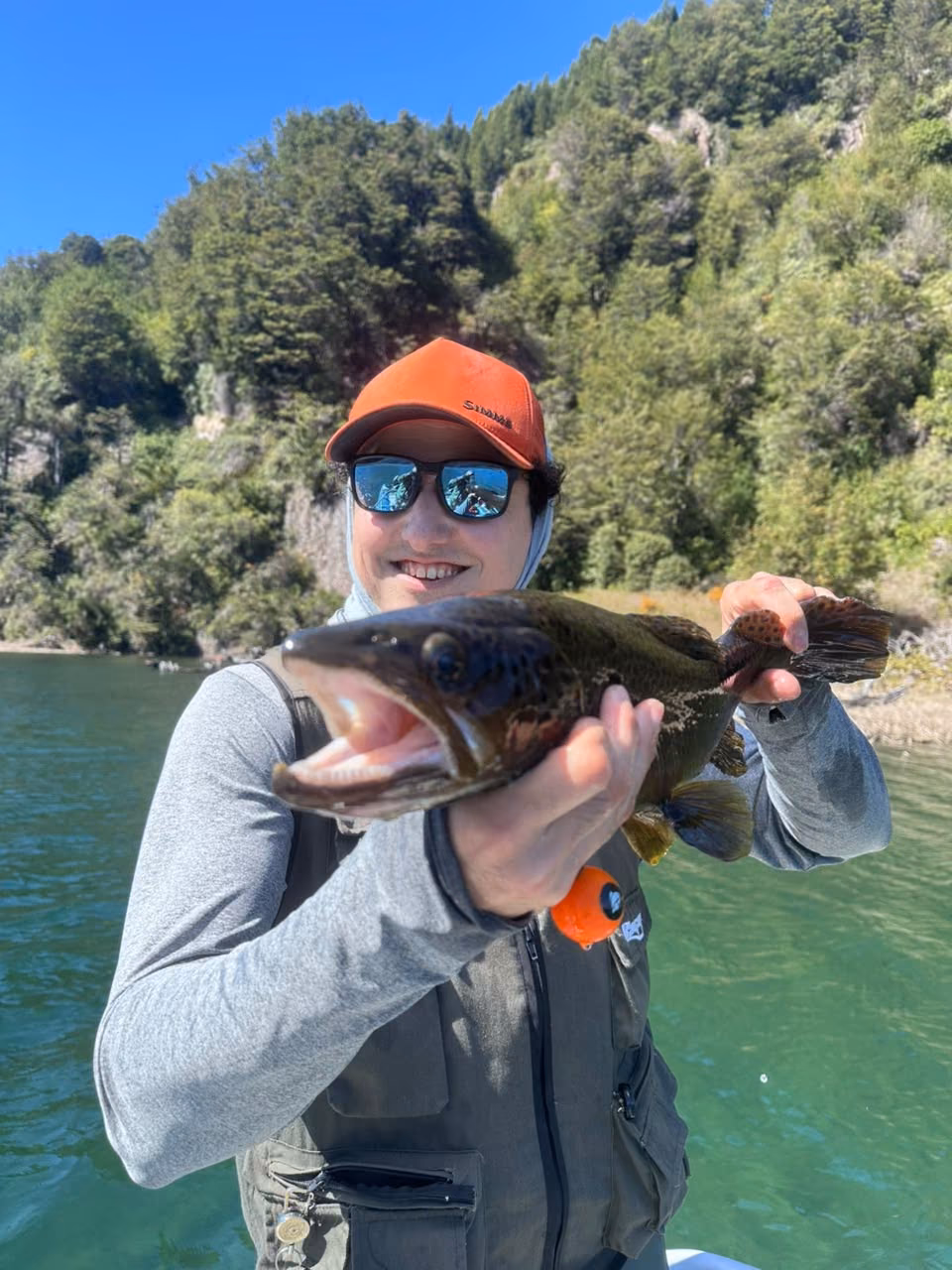 An angler in an orange cap holds a dark, open-mouthed brown trout from a boat on a clear lake near Coyhaique, Chilean Patagonia