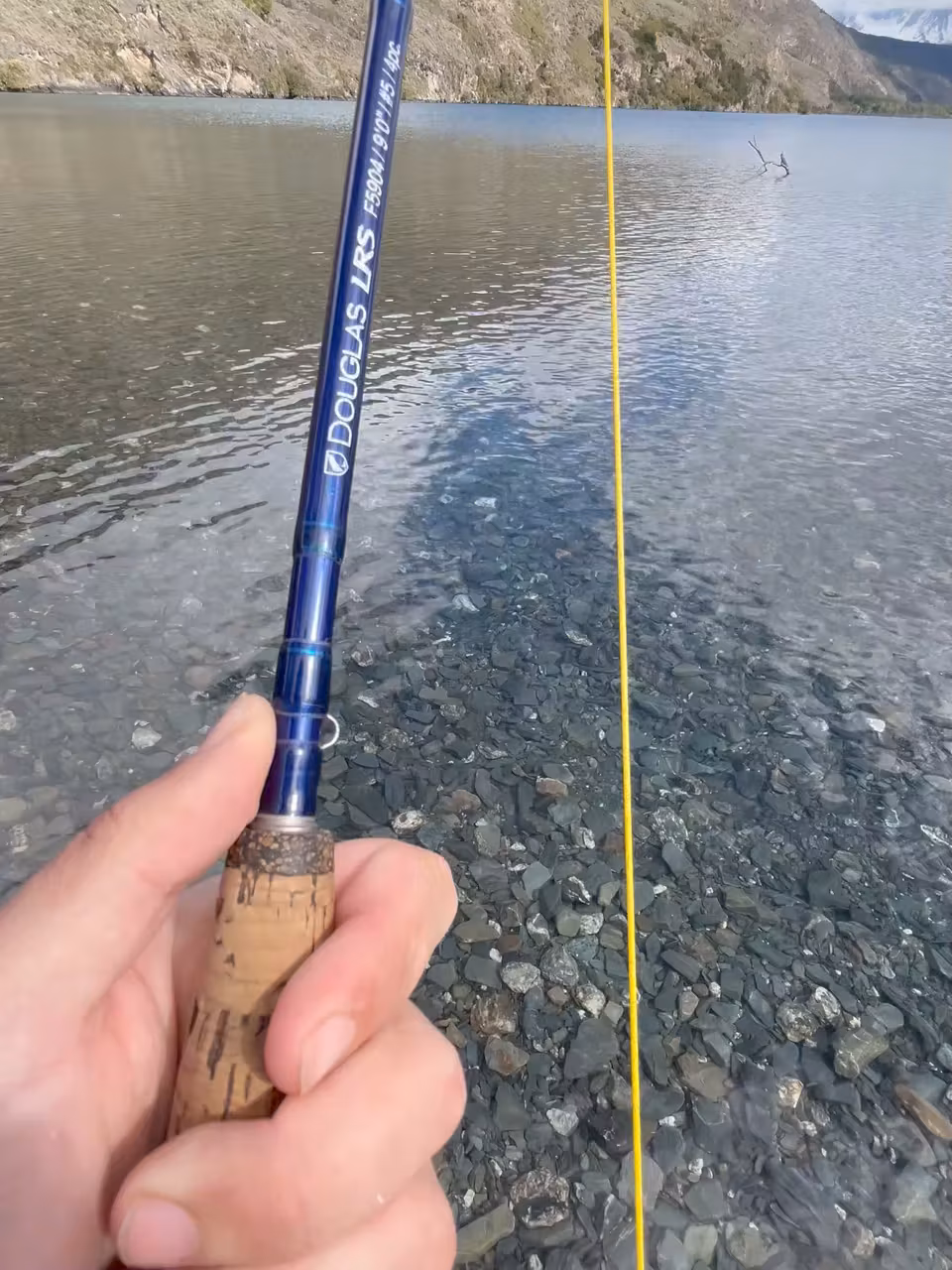 First-person view of a fly rod and a yellow fly line over a pale stone shelf of clear shallow water that falls away to deep water, with a hillside and a lake behind, Chilean Patagonia