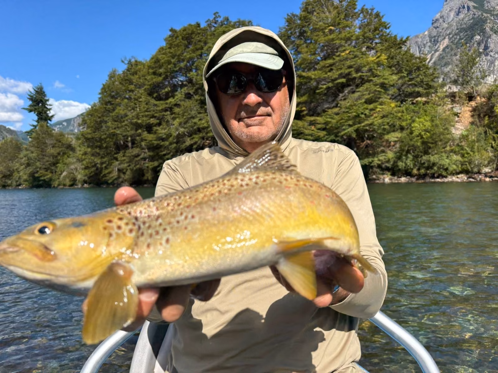 An angler in a hooded sun shirt, green cap and dark sunglasses holds a golden brown trout with dark and red spots over clear river water, a forested bank and rocky mountain behind, Coyhaique, Chilean Patagonia
