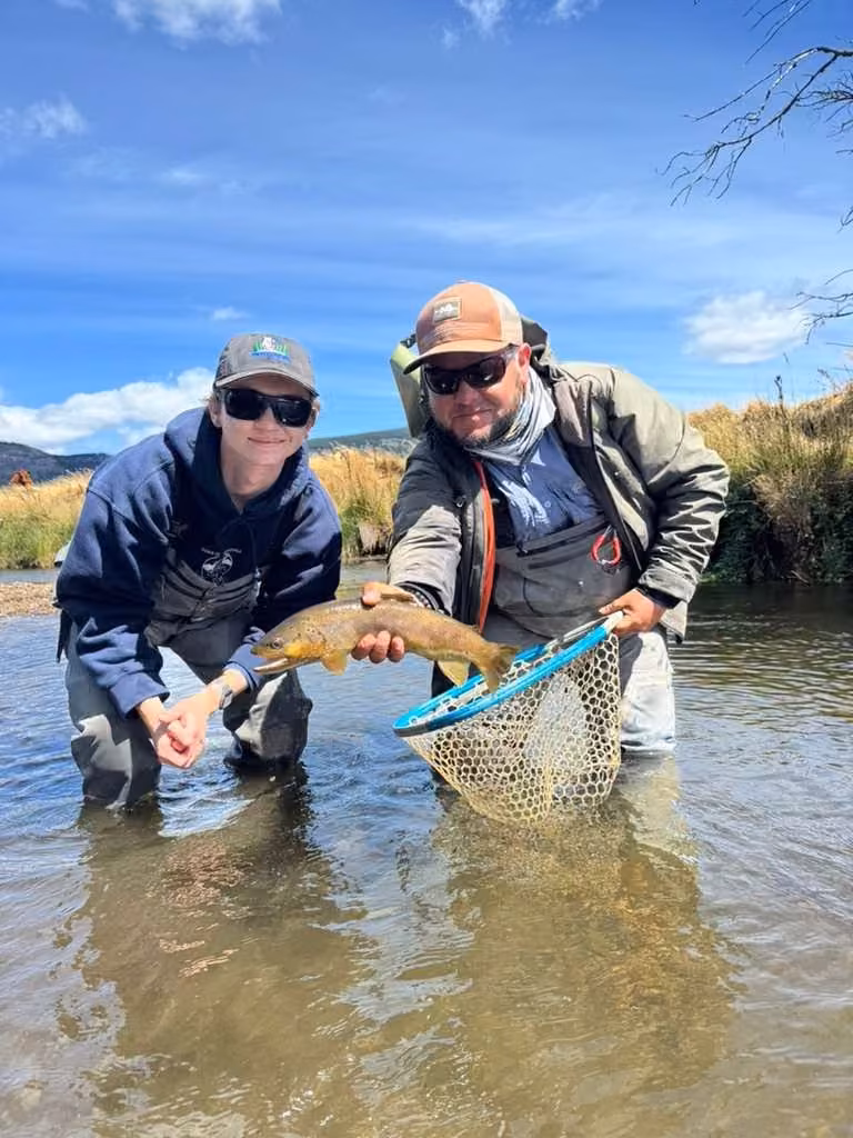 Two people stand in shallow river water under a blue sky, one in an orange cap holding a brown trout over a landing net and one in a navy hoodie beside, Ñirehuao River, Coyhaique, Chilean Patagonia