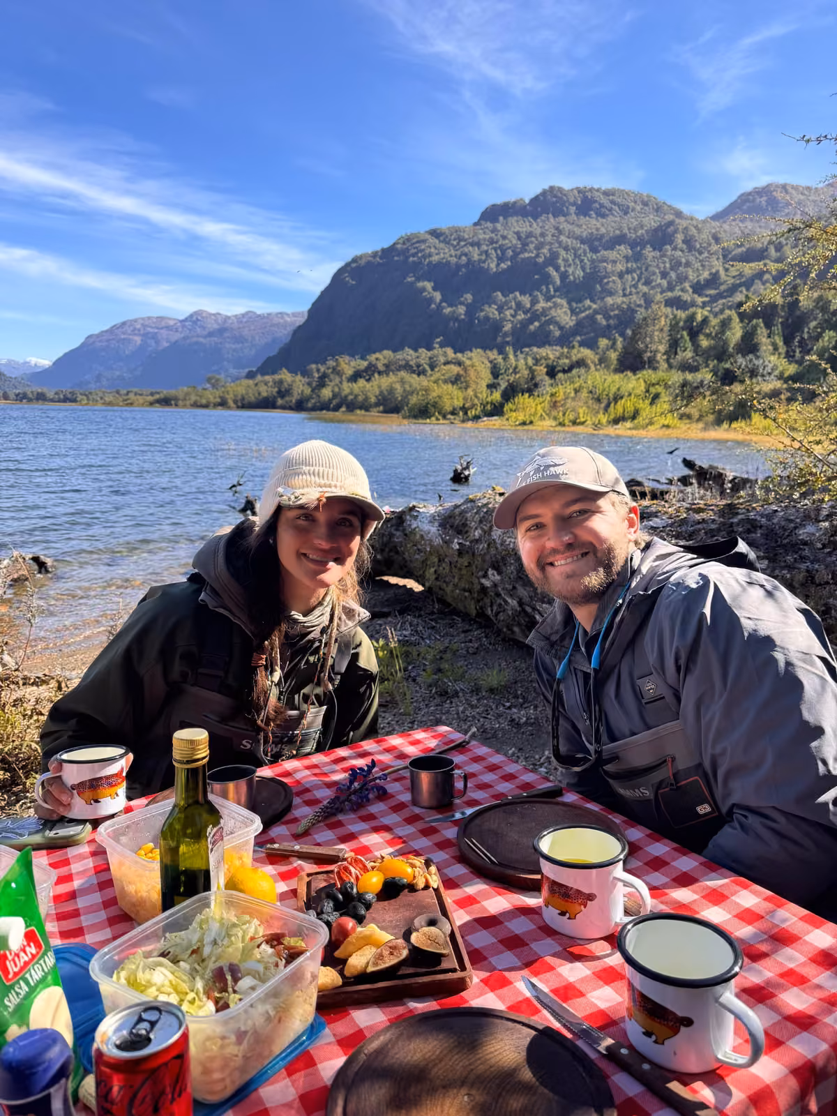 Two smiling anglers in waders sit at a table with a red-and-white checked cloth, enamel mugs and salad in plastic containers beside a lake, a forested mountain behind, Coyhaique, Chilean Patagonia