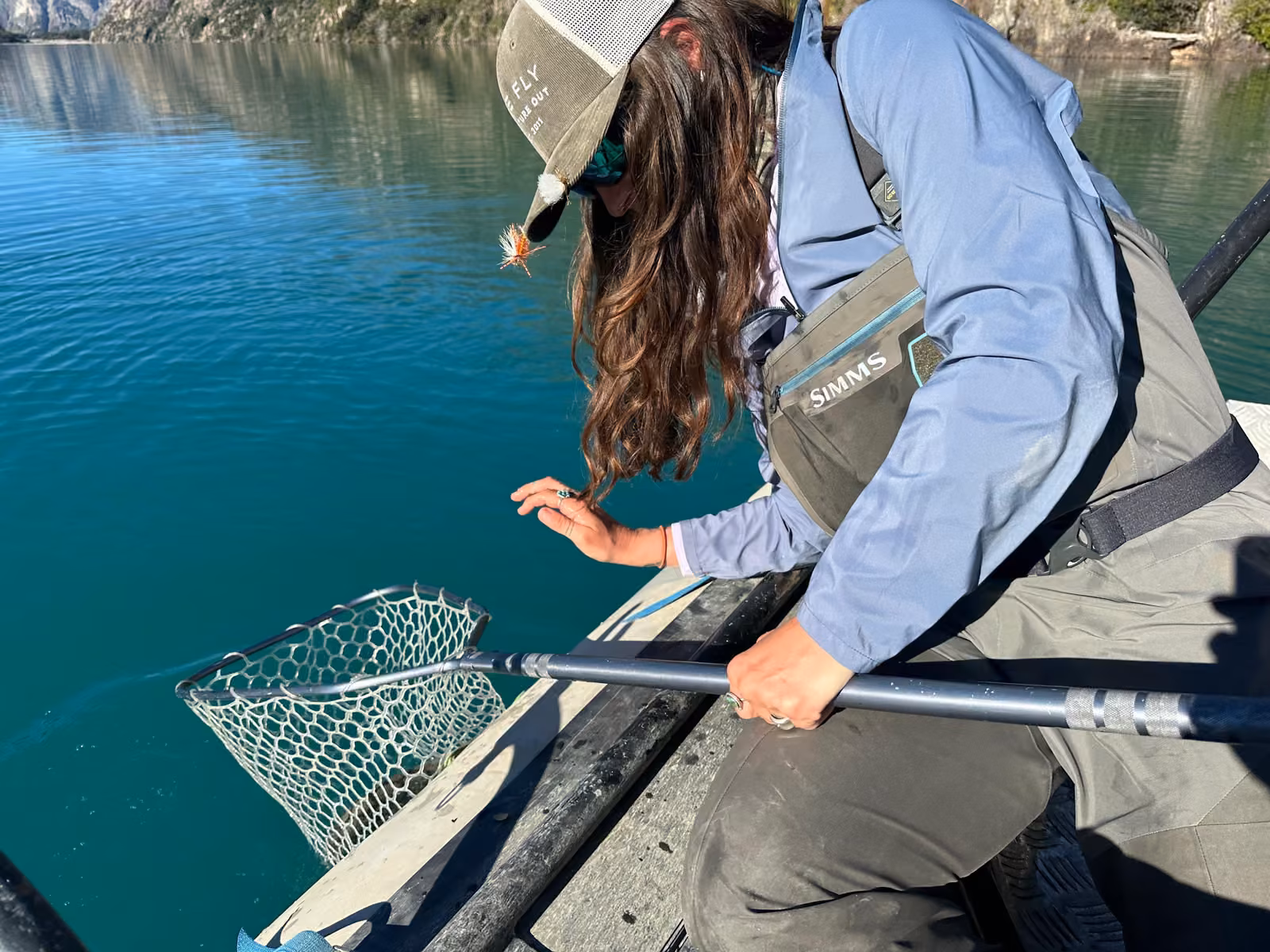 An angler in waders leans over the side of a boat and holds a landing net in the turquoise water of a lake, Coyhaique, Chilean Patagonia