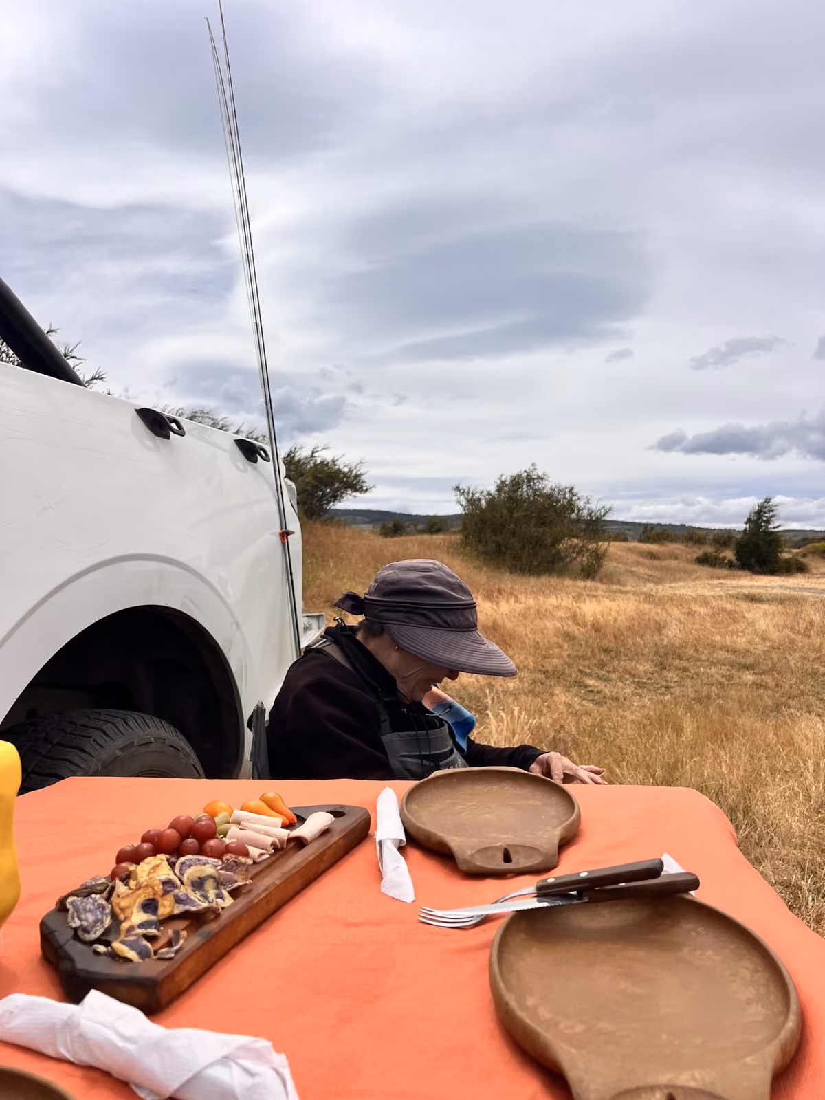 A table with an orange cloth beside a white truck, set with a wooden snack board, a knife and fork and wooden plates, an angler in a hat seated at the truck, Coyhaique, Chilean Patagonia