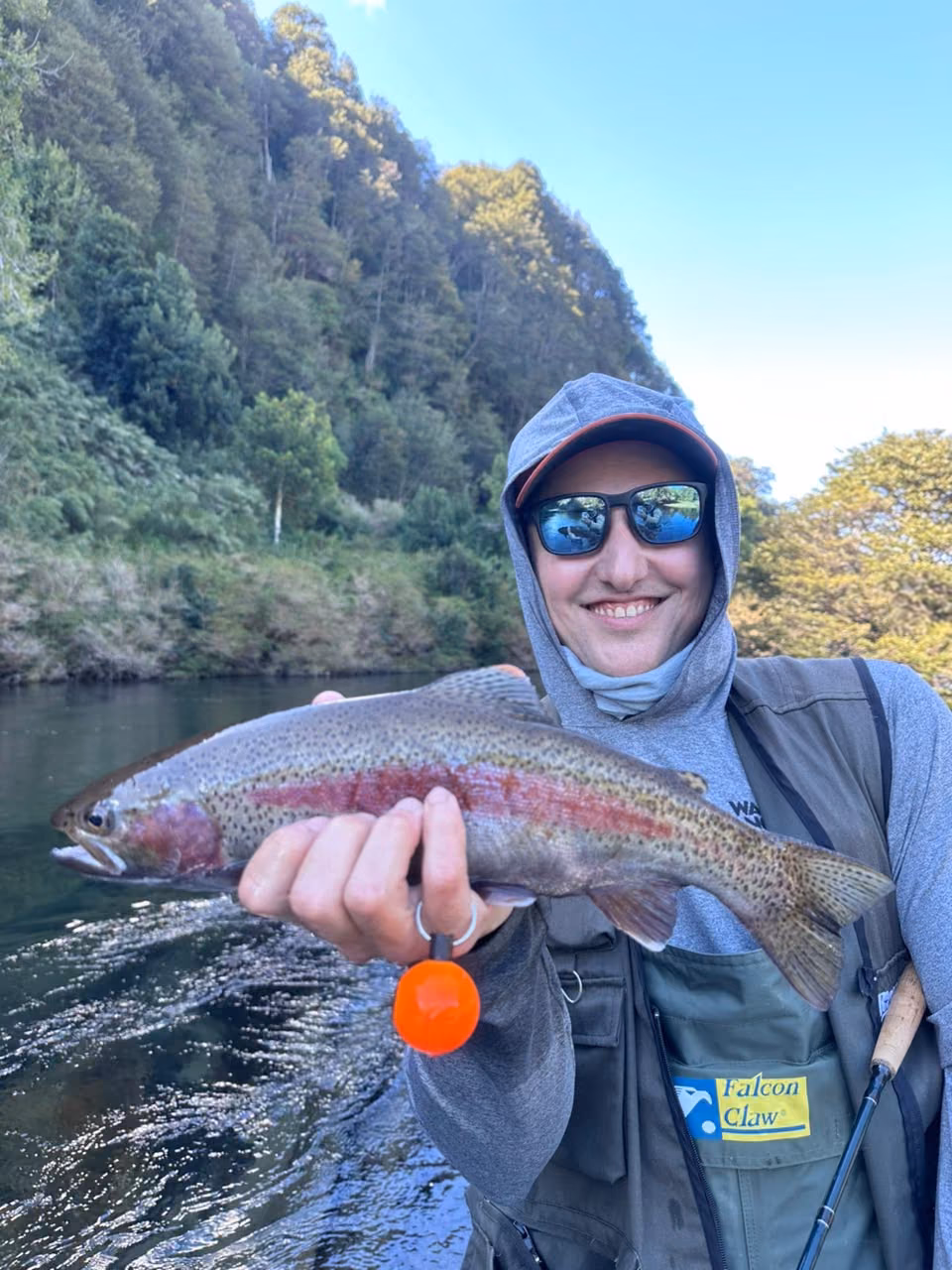 An angler in a hood and polarized sunglasses holds a rainbow trout with a red band along its flank, Coyhaique, Chilean Patagonia