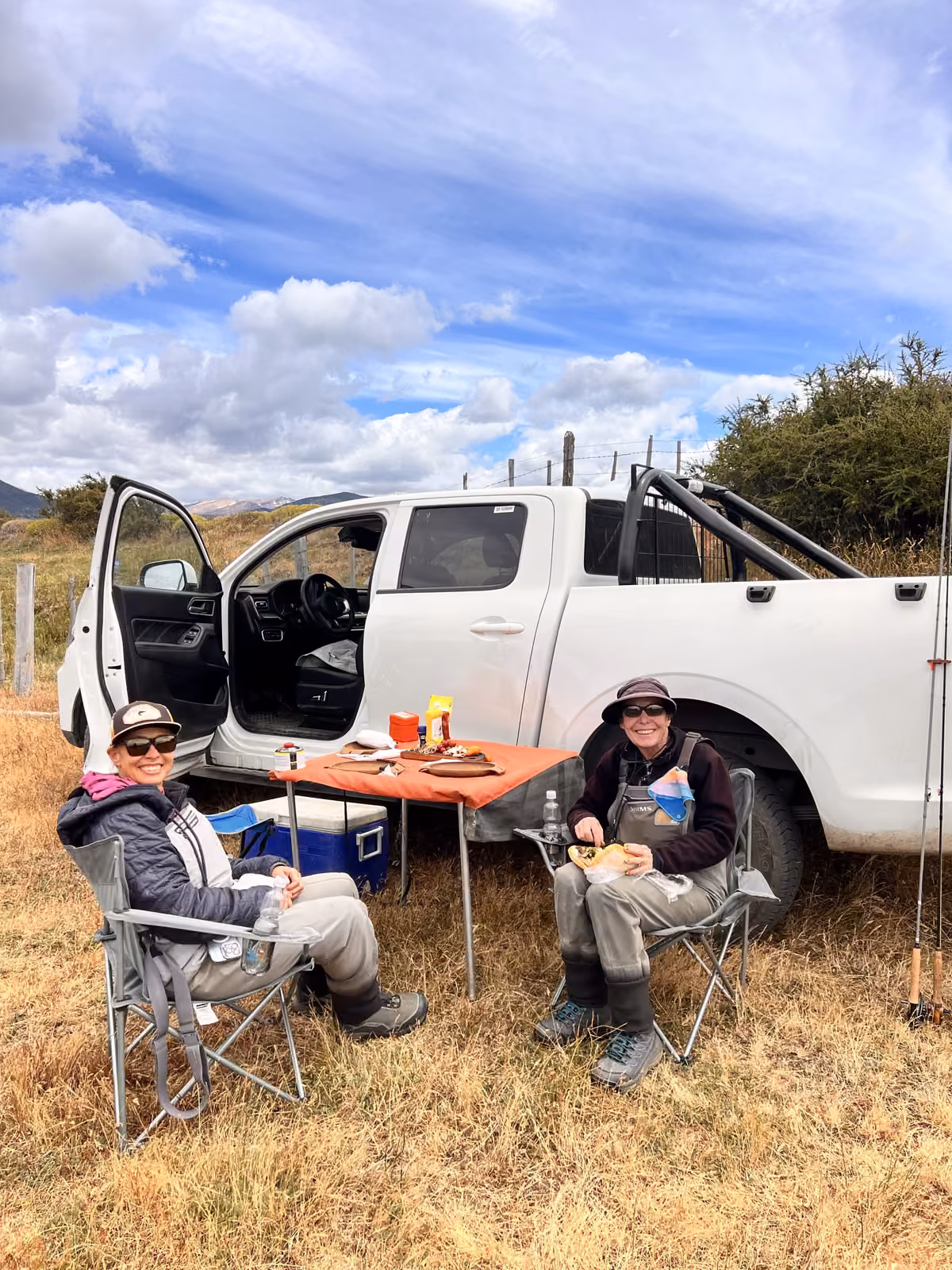 Two anglers in waders sit in camp chairs at a table with an orange cloth and a cooler, beside a white pickup truck with its door open in dry golden grassland, Coyhaique, Chilean Patagonia
