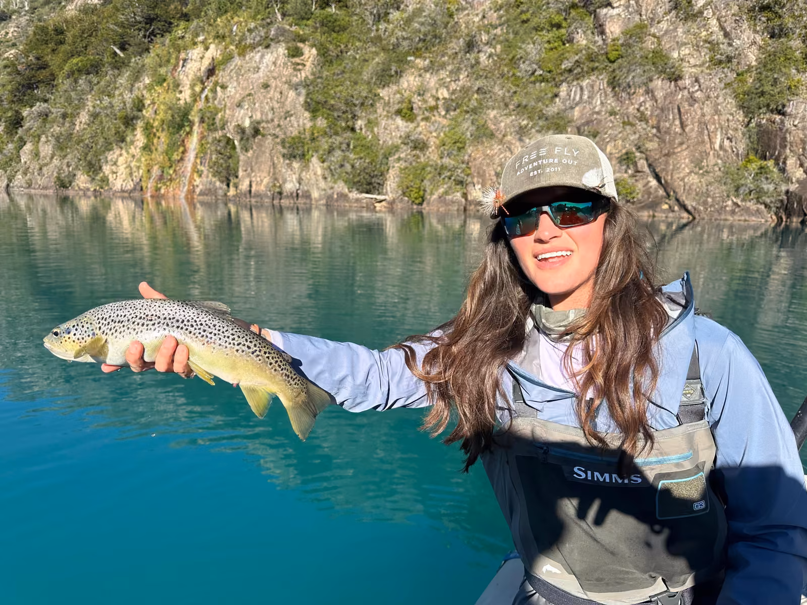 A smiling woman angler in a cap and waders holds a spotted brown trout out over the turquoise water of a lake, a steep rock wall behind, Coyhaique, Chilean Patagonia