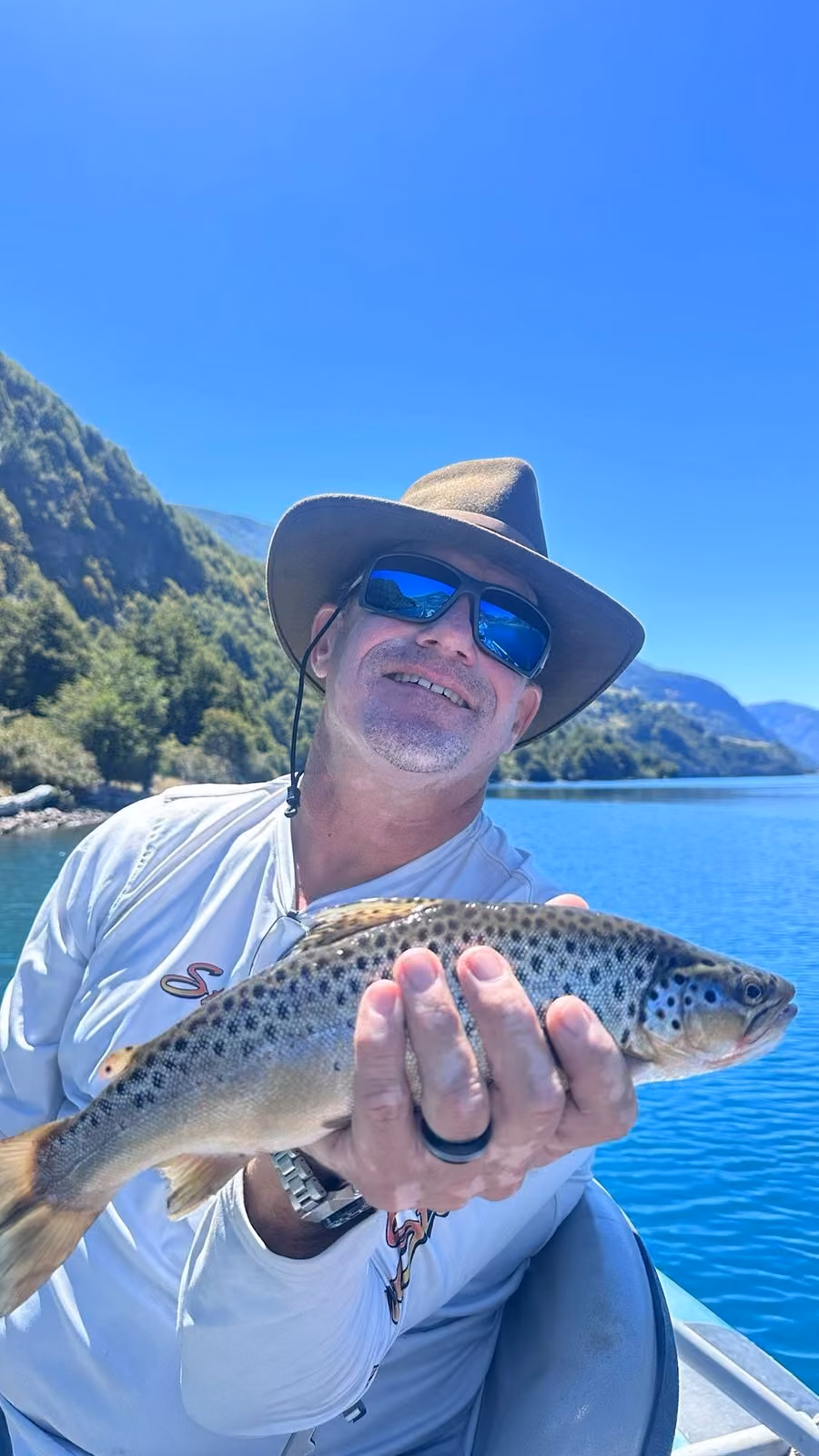 An angler in a wide-brim hat and blue mirrored sunglasses holds a dark-spotted brown trout in a boat on a deep blue lake, a forested mountainside behind, Coyhaique, Chilean Patagonia