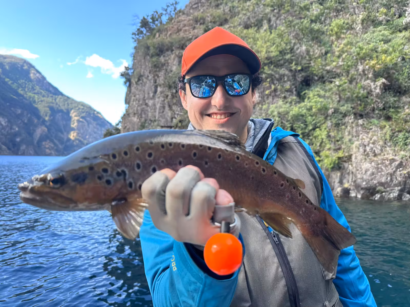 A smiling angler in an orange cap and polarized sunglasses holds a dark, heavily spotted brown trout beside a vegetated rock wall on Lago Azul, Coyhaique, Chilean Patagonia