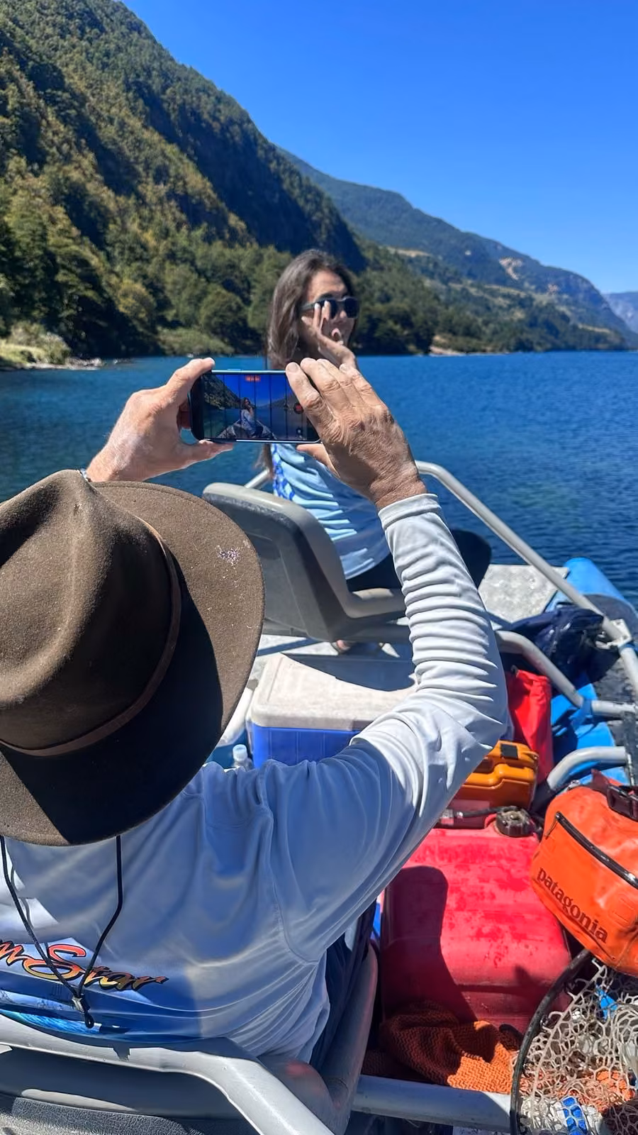 An angler in a brown wide-brim hat holds up a phone to photograph a woman in sunglasses seated in a boat on a blue lake, forested slopes behind, Coyhaique, Chilean Patagonia
