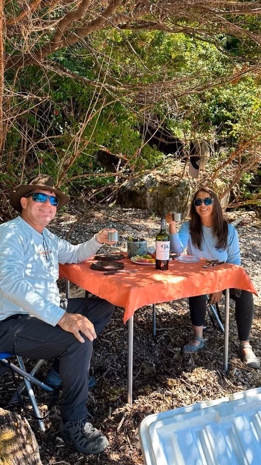 A man in a wide-brim hat and a woman in sunglasses raise metal cups at a table with an orange cloth on a pebbly shore, with plates, cutlery and a bottle, Coyhaique, Chilean Patagonia