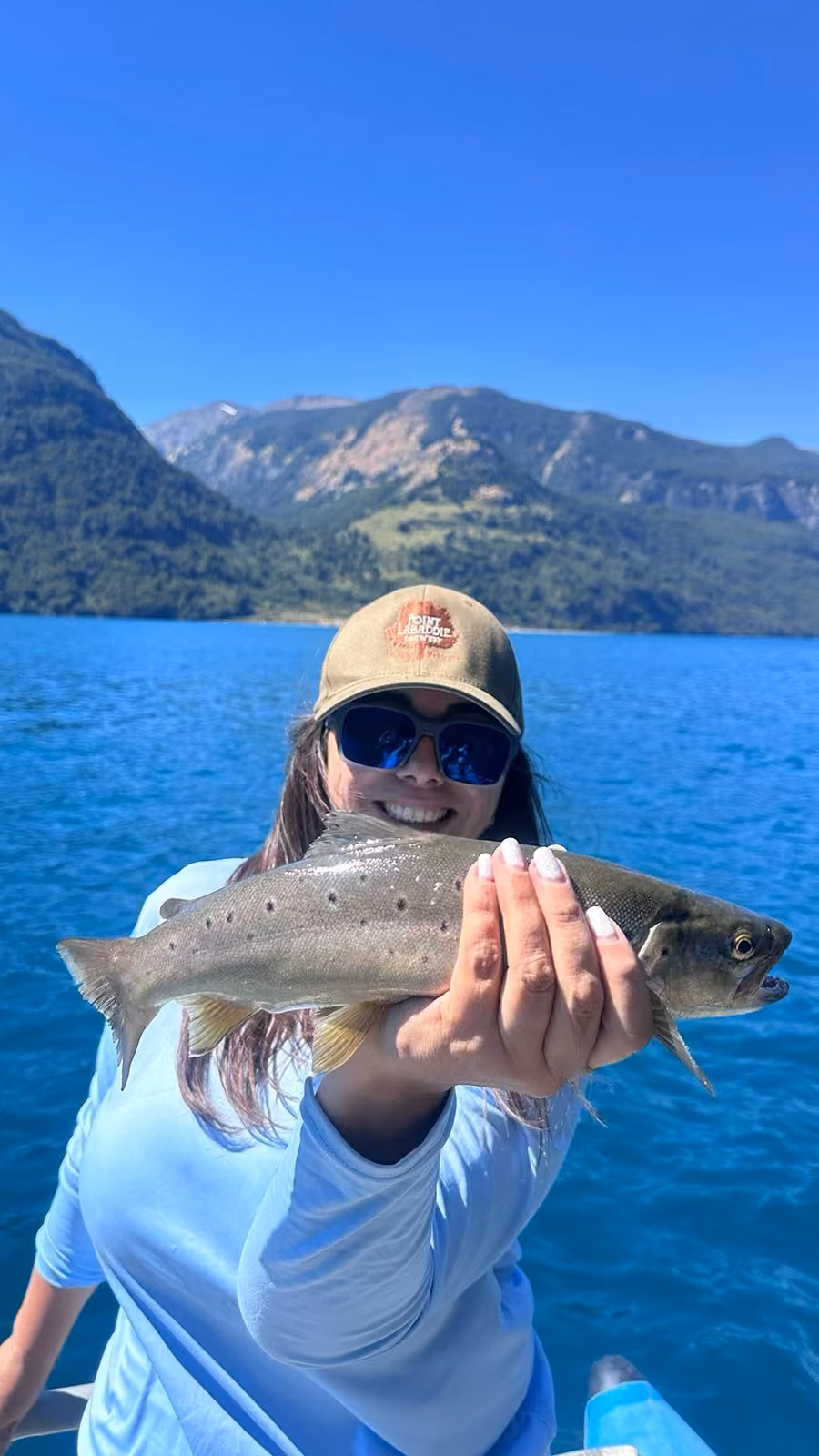 A smiling angler in an olive cap and blue mirrored sunglasses holds a silvery brown trout with scattered dark spots over deep blue lake water, green mountains behind, Coyhaique, Chilean Patagonia