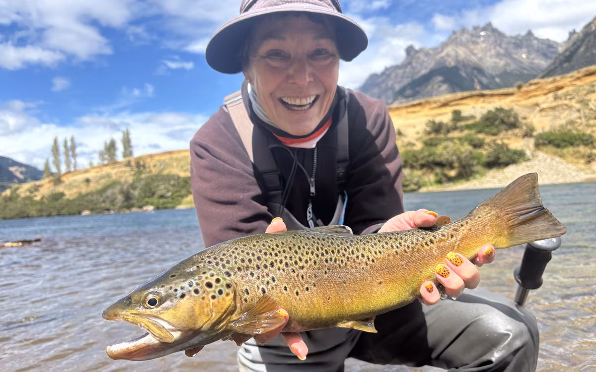 A smiling angler in a lavender bucket hat holds a brown trout in shallow water, with dry hills and a rocky peak behind, Coyhaique, Chilean Patagonia