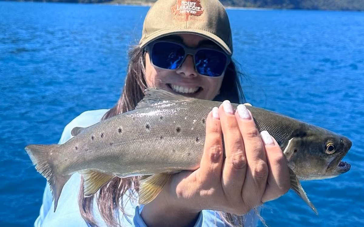 A smiling angler in an olive cap and blue mirrored sunglasses holds a brown trout over deep blue lake water, Coyhaique, Chilean Patagonia