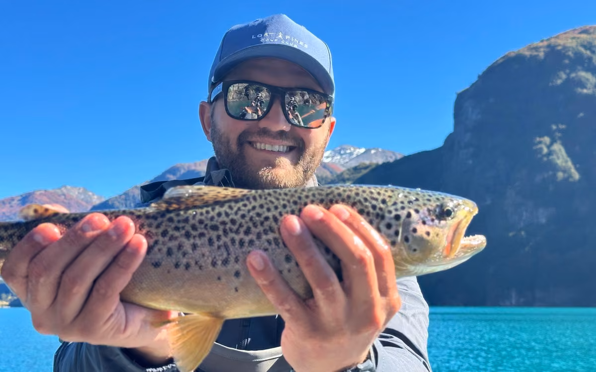 A smiling angler in a blue cap and mirrored sunglasses holds a spotted brown trout, a turquoise lake and a rock wall behind, Coyhaique, Chilean Patagonia